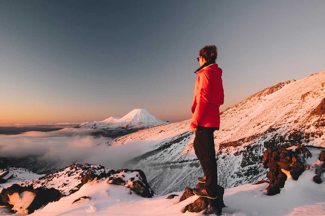 Hiker in winter jacket standing on snowy mountain, admiring view of snow-capped peaks and clouds at sunrise in Tongariro National Park, New Zealand