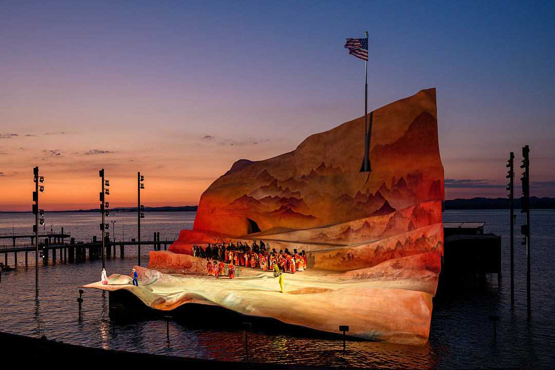 Open-air opera performance on a floating stage at sunset with a dramatic backdrop and calm waters