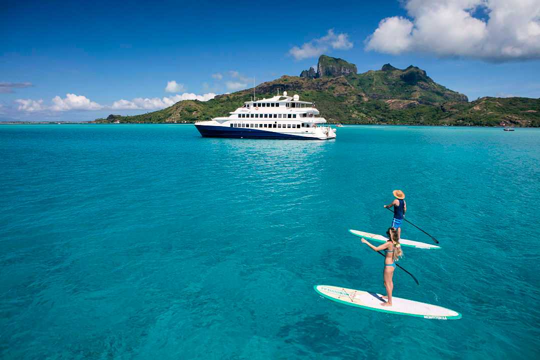 Couple paddleboarding on turquoise water in Bora Bora with a luxury yacht and tropical island backdrop