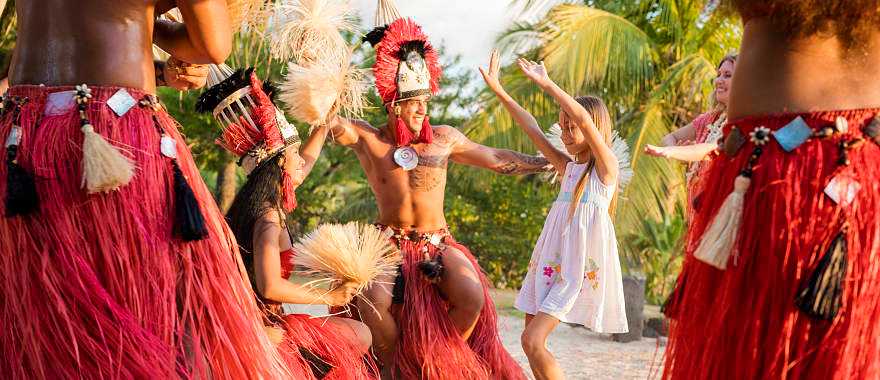 Young girl and her mother with Tahitian men and women in traditional attire performing a cultural dance surrounded by lush greenery and a serene ocean backdrop in Tahiti